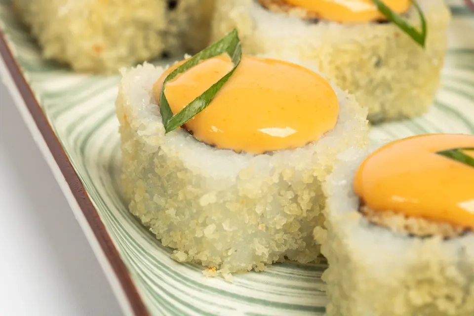 Close-up shot of sushi rolls, each topped with a creamy yellow-orange sauce and garnished with a thin slice of green onion. The sushi rolls have a crispy exterior and are served on a green and white patterned plate.