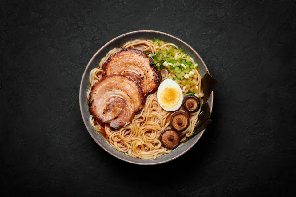 A bowl of ramen sits on a dark, textured surface. The dish includes slices of meat, half a boiled egg, sliced mushrooms, green onions, and noodles in a savory broth. Seaweed is also visible on the edge of the bowl.