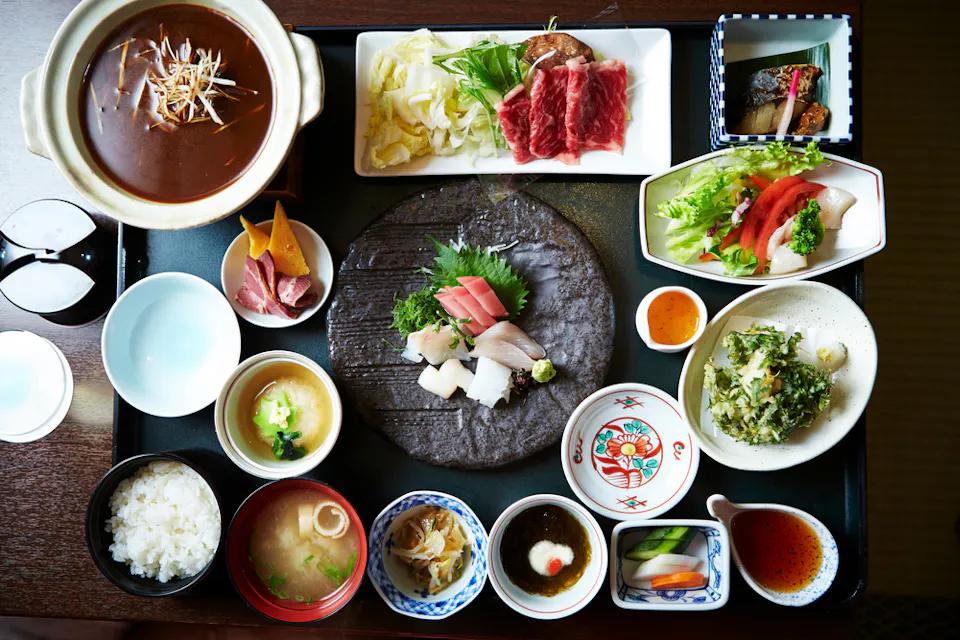 A traditional Japanese meal served on a tray, including various dishes such as sashimi, salad, rice, miso soup, pickled vegetables, and cooked fish. The meal appears vibrant and neatly presented in separate bowls and plates, showcasing diverse textures and colors.