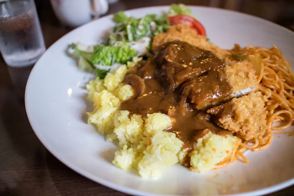 A white plate with a portion of breaded fried meat topped with brown gravy, served with yellow rice, spaghetti in tomato sauce, and a side salad with lettuce and tomato. A glass of water is visible in the background.