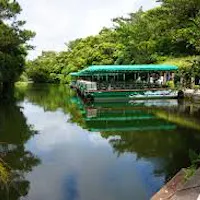 Bios no Oka (Bios on the Hill) A green boat with a canopy is docked on a calm river surrounded by lush green trees, with reflections visible in the water under a partly cloudy sky.