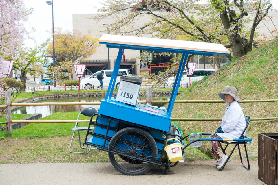 A woman wearing a hat and white jacket is sitting on a chair next to a blue street vendor cart with a white roof. The cart, priced at 150 yen, is parked on a pathway in a park with blooming cherry blossom trees and a pond in the background.