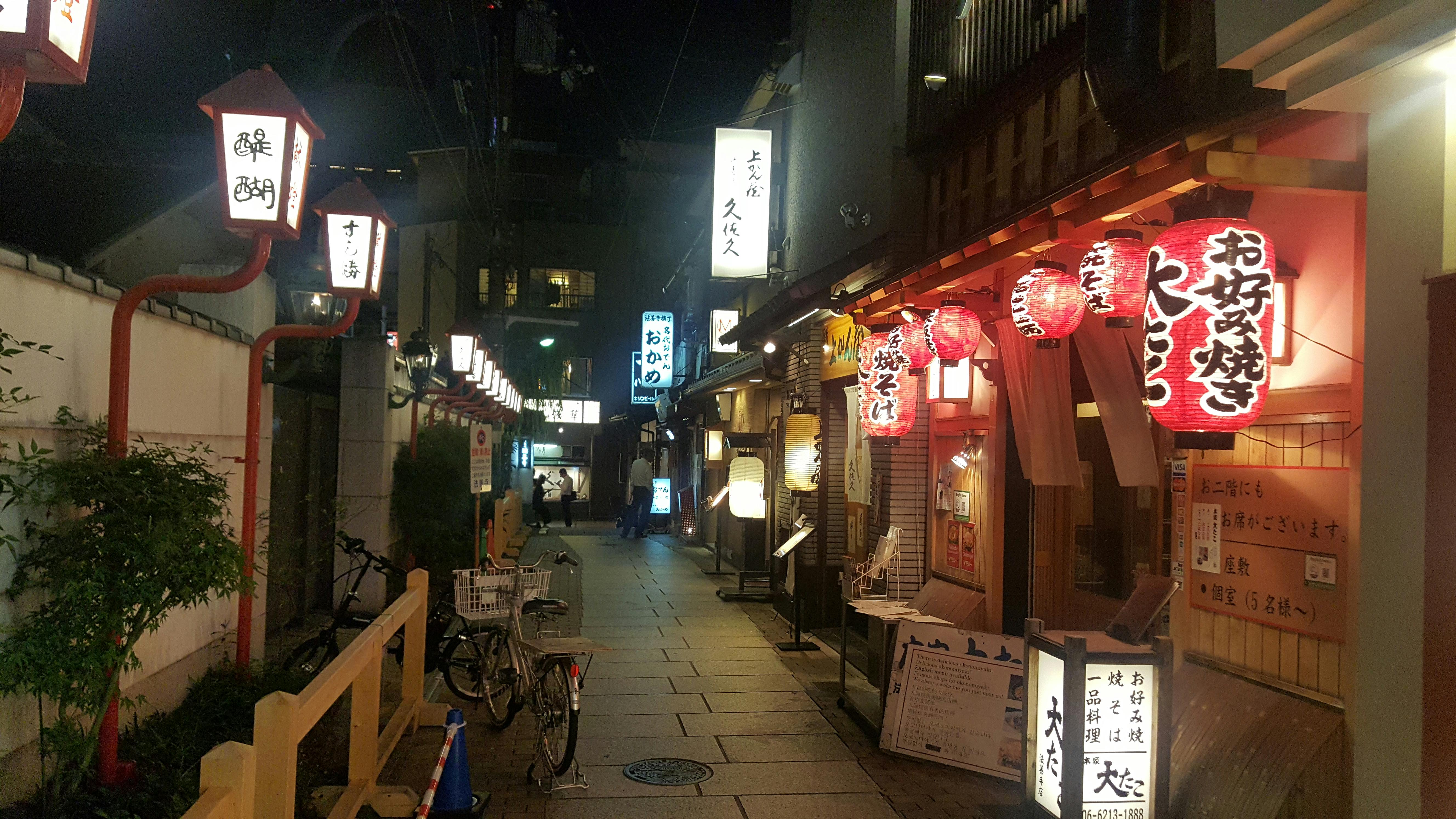 A narrow, lantern-lit street in Japan at night, lined with traditional wooden buildings and glowing signs featuring Japanese characters. Bicycles are parked along the path, and red paper lanterns hang outside restaurants.
