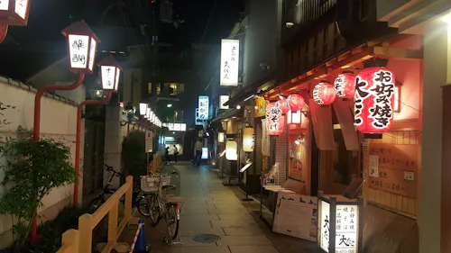 A narrow, lantern-lit street in Japan at night, lined with traditional wooden buildings and glowing signs featuring Japanese characters. Bicycles are parked along the path, and red paper lanterns hang outside restaurants.