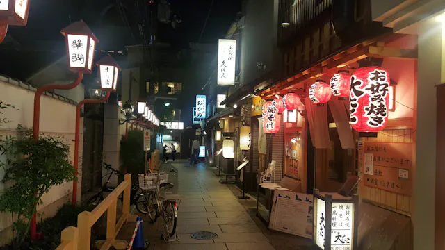 A narrow, lantern-lit street in Japan at night, lined with traditional wooden buildings and glowing signs featuring Japanese characters. Bicycles are parked along the path, and red paper lanterns hang outside restaurants.