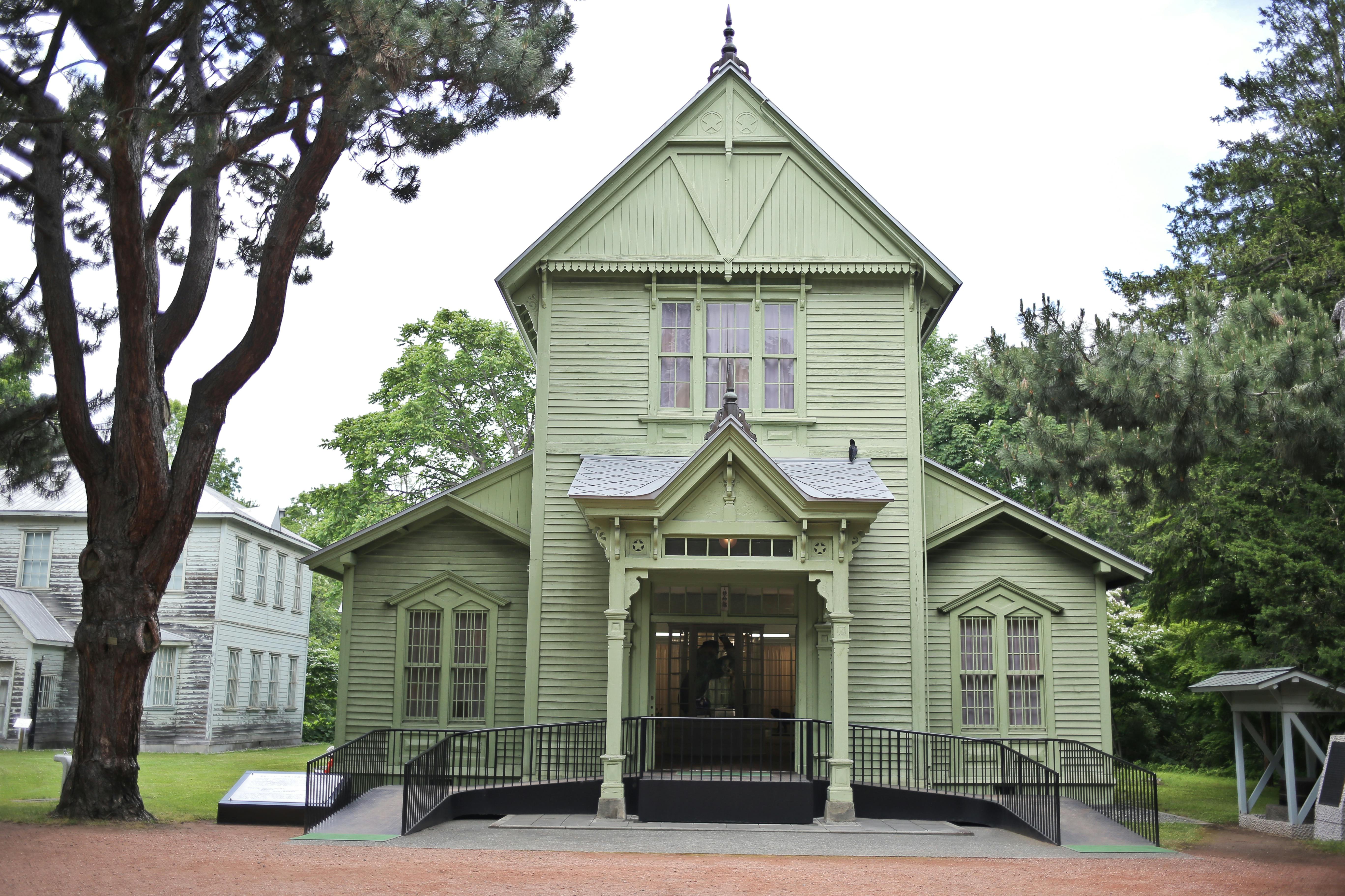 A pale green, wooden, historic building with ornate trim and pointed roof stands among trees. The entrance features a covered porch with decorative columns and a ramp leading to the doorway.