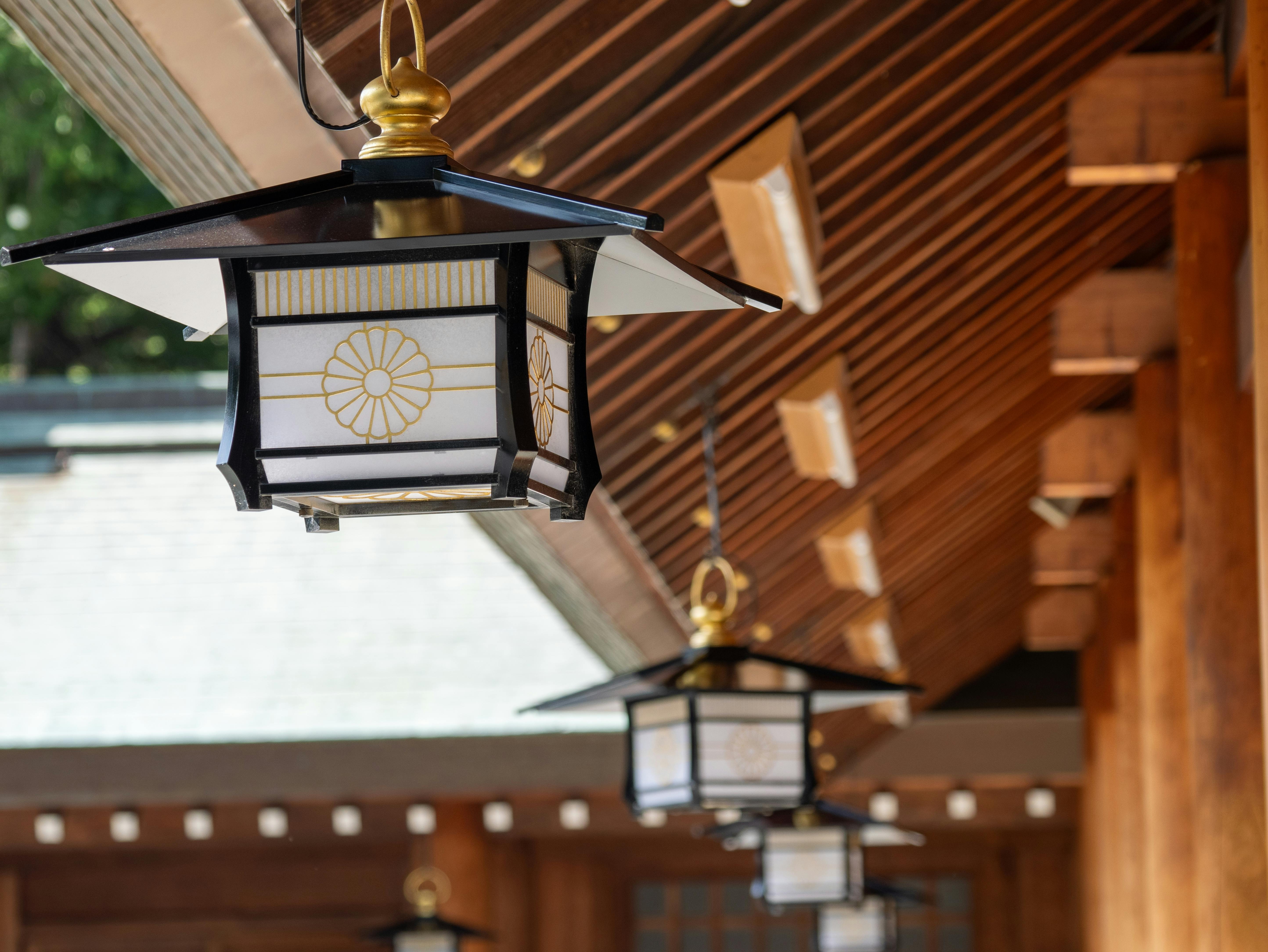 Traditional Japanese lanterns with gold chrysanthemum emblems hang under the eaves of a wooden building, featuring angled roof beams and natural light highlighting the structure.