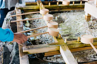 Close-up of a person’s hand holding a wooden ladle over a stone basin filled with water and white pebbles, at a traditional Japanese purification fountain. Several other ladles rest on a wooden frame above the basin.