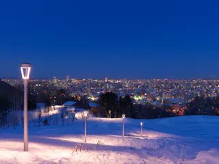 Snow-covered hill with lit lampposts overlooking a brightly illuminated cityscape at dusk. The sky is deep blue, and buildings stretch across the horizon, creating a vibrant urban scene.