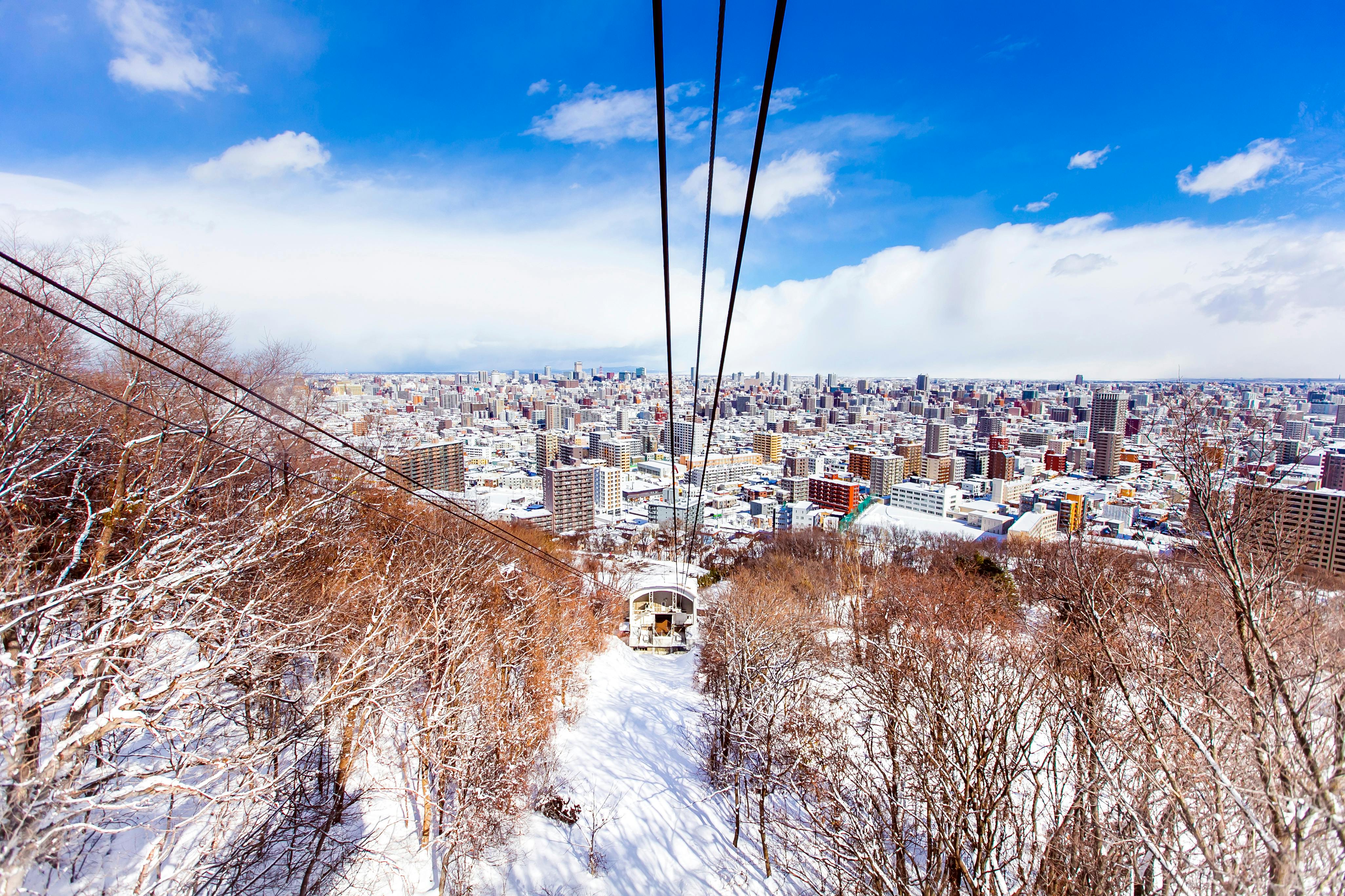 A cable car travels above a snowy forest toward a cityscape under a bright blue sky, with tall buildings and snow-covered trees visible below.