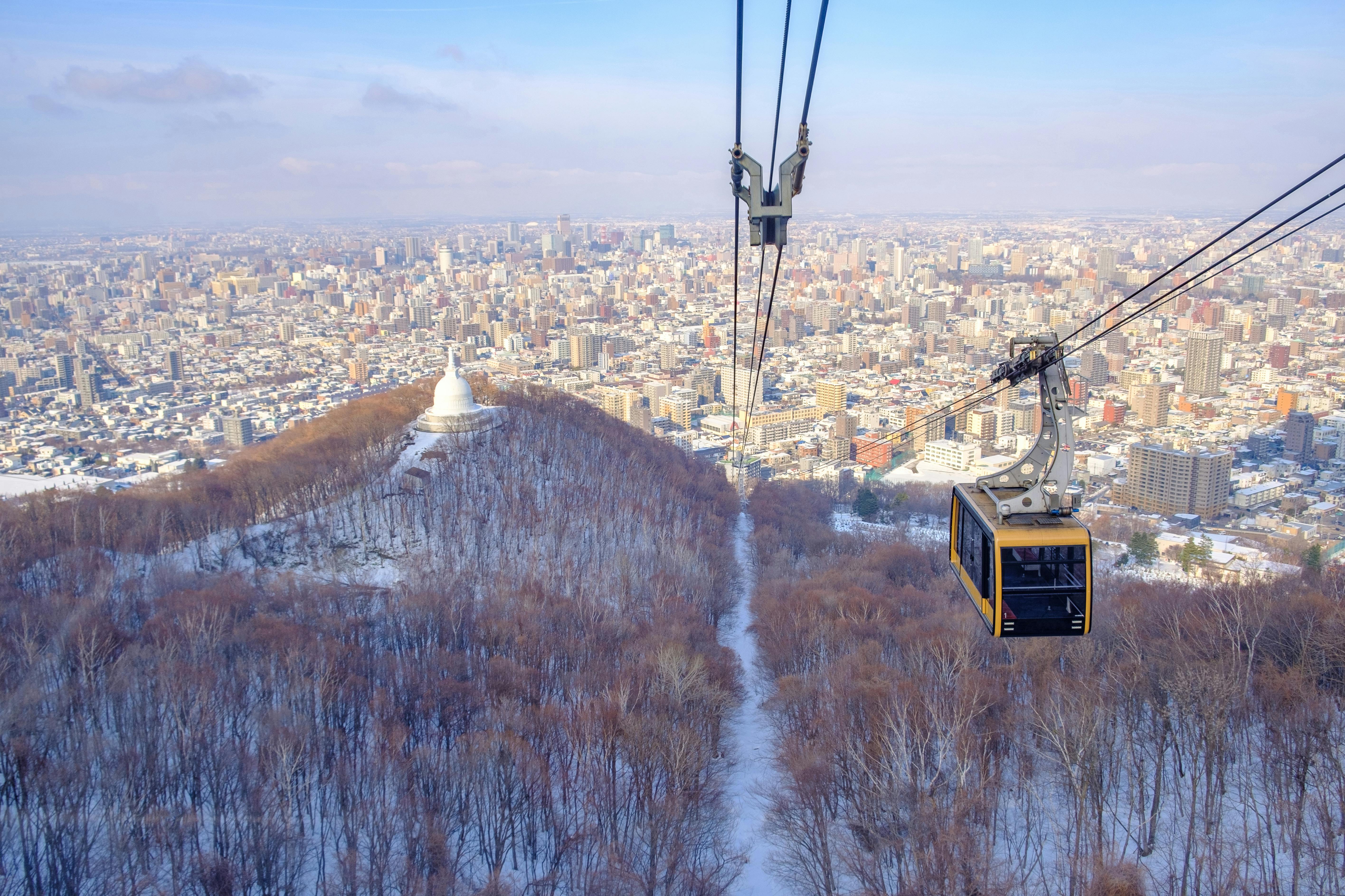 A yellow cable car ascends over a snowy, tree-covered hill toward a cityscape, with a large white structure resembling a stupa visible among the trees under a clear blue sky.