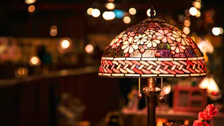 A stained glass table lamp with a floral pattern glows warmly in a dimly lit room, creating a cozy and inviting ambiance. The background is softly blurred with twinkling lights.
