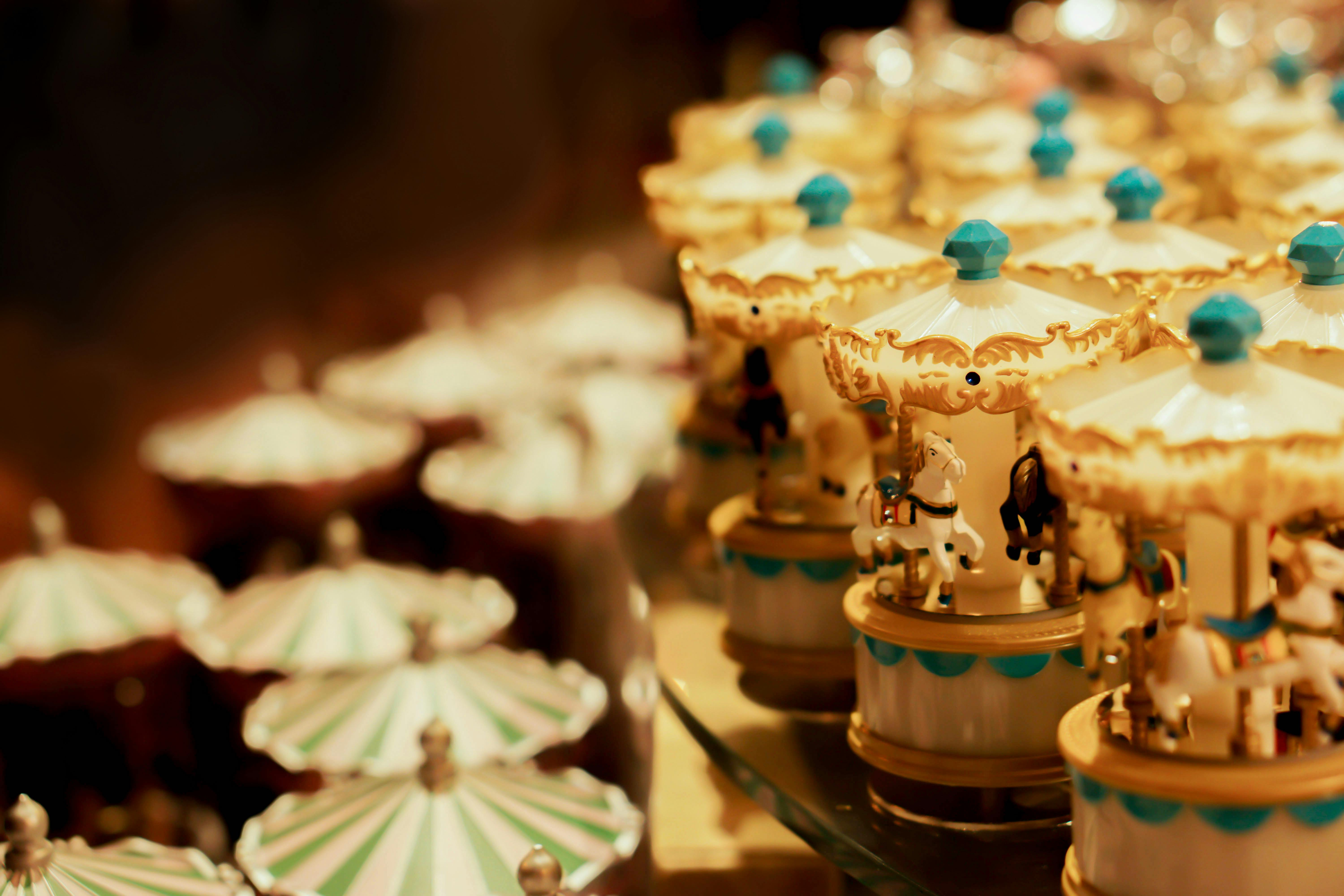 A close-up of miniature carousel music boxes in soft, warm lighting. The carousels are decorated with white and blue colors, and small horses are visible under the canopies. The background is blurred.