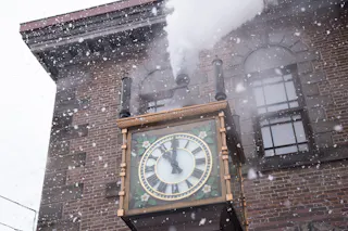 A steam clock on a brick building emits steam as snow falls. The clock shows 12:18, and the scene is wintry with snowflakes in the air and accumulated on the clock and windowsills.