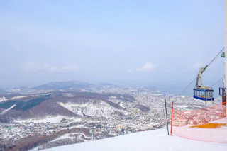 A blue cable car ascends over a snowy mountain slope, overlooking a town and forested hills with the sea and distant mountains visible under a pale blue sky.