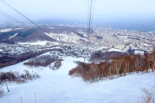 A snowy ski slope with cable car lines overhead descends toward a town nestled among hills and trees, with a body of water and mountains visible in the background under a hazy sky.