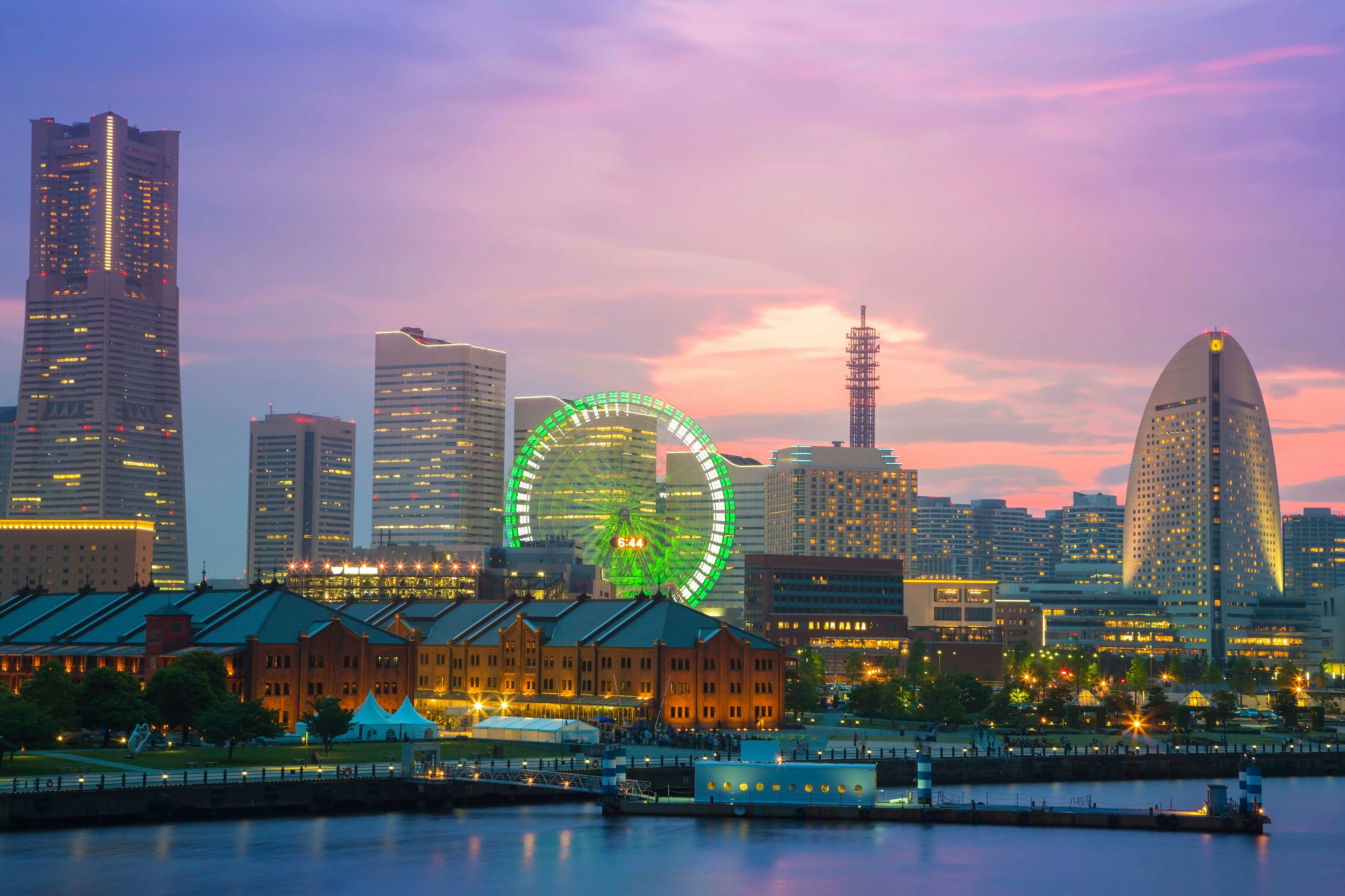 City skyline at dusk featuring tall modern buildings, a glowing green Ferris wheel, and a curved white structure by the waterfront, with colorful lights reflecting on the water and a purple-pink sky in the background.