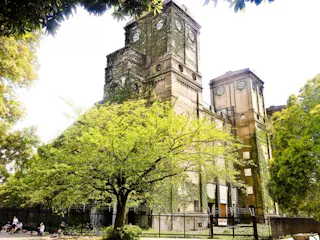 An old, ivy-covered building with two clock towers stands behind a black iron fence, surrounded by lush green trees on a sunny day. A few people and bicycles are visible near the entrance.