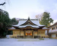 A traditional Japanese Shinto shrine with a curved, ornate roof and wooden architecture, surrounded by trees and lit softly, photographed from the front at dusk.