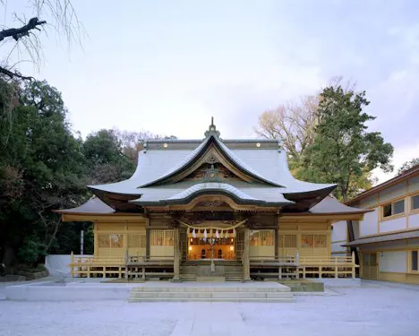 A traditional Japanese Shinto shrine with a curved, ornate roof and wooden architecture, surrounded by trees and lit softly, photographed from the front at dusk.