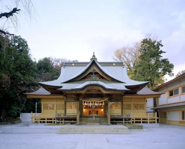 A traditional Japanese Shinto shrine with a curved, ornate roof and wooden architecture, surrounded by trees and lit softly, photographed from the front at dusk.