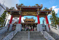 A traditional Chinese temple with ornate red columns, gold decorations, and dragon sculptures atop the roof, seen from the base of a staircase with people walking up towards the entrance under a blue sky.