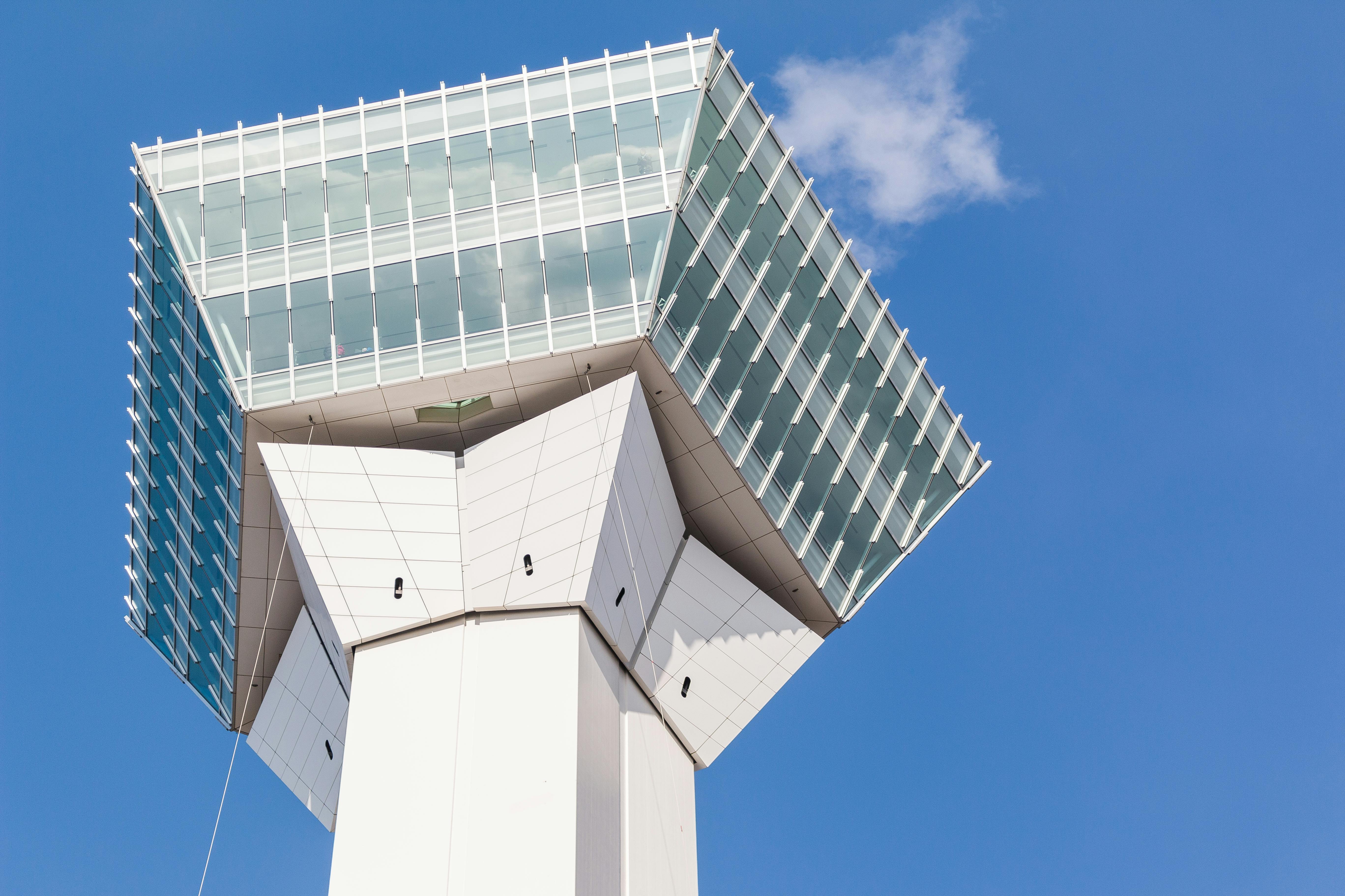 A modern, white observation or control tower with large, angled glass windows stands against a clear blue sky with a small white cloud.