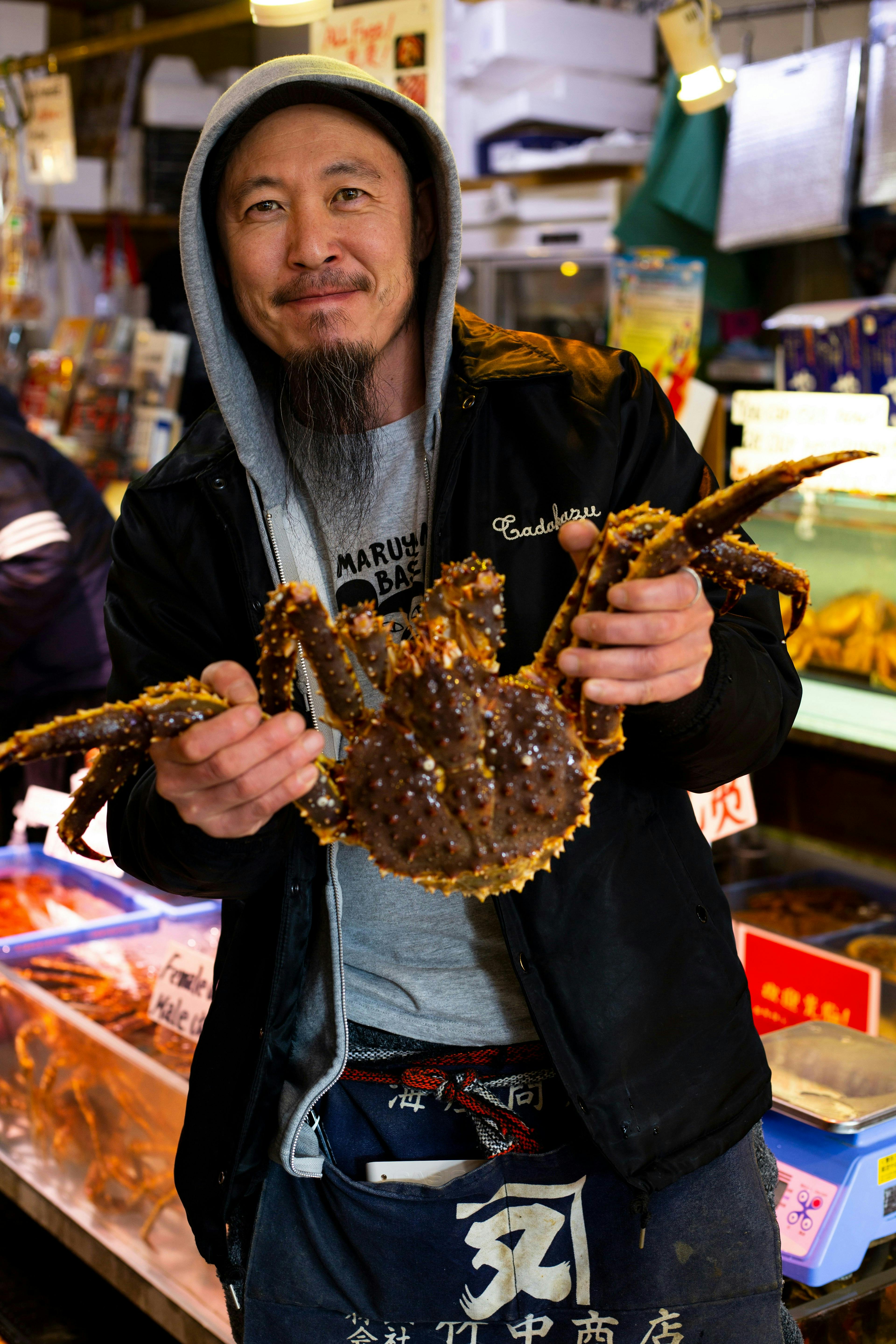 A man wearing a hoodie and jacket smiles while holding a large spiky crab with outstretched legs at a seafood market. Tanks and trays of seafood are visible in the background.