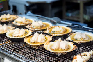 Scallops in their shells are being grilled on a metal grate, with pieces of raw scallop meat placed on top, likely at a seafood market or food stall.