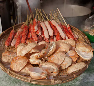 A woven basket filled with empty crab shells in the foreground and several skewers of crab meat stacked in the background, with a steaming pot partially visible behind them.