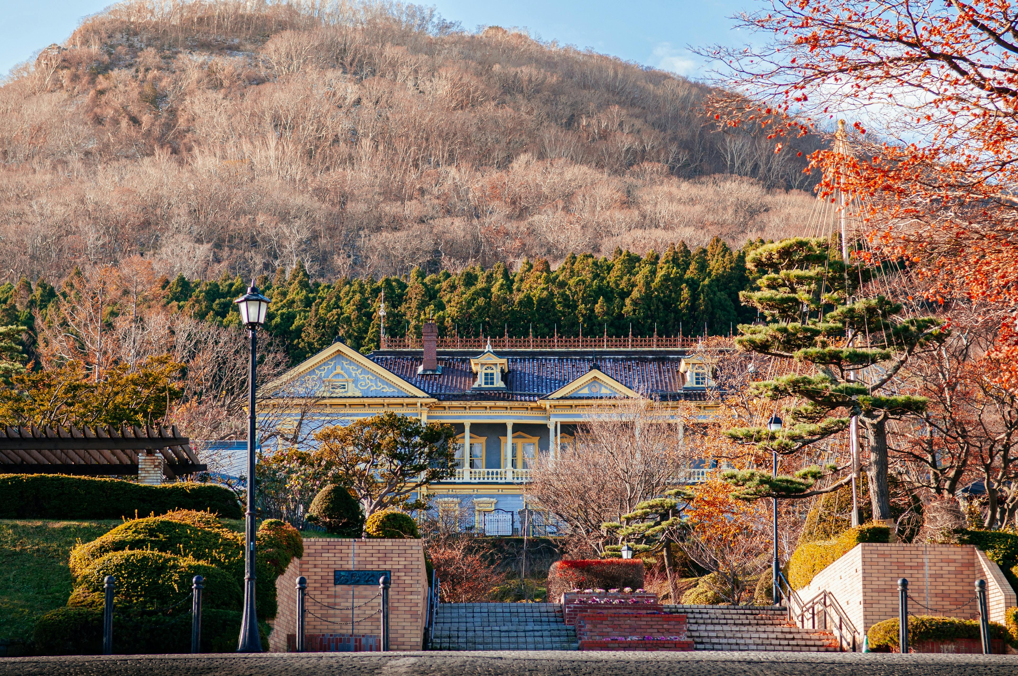 A yellow, Western-style building stands amid manicured gardens, with stone steps leading up to it. Behind, a forested hillside with bare trees rises under a clear sky. Autumn foliage frames the scene.