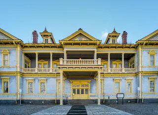 A two-story historic building with pale blue walls and bright yellow trim, ornate balconies, and a symmetrical design, set under a clear blue sky. The entrance features double doors and decorative columns.