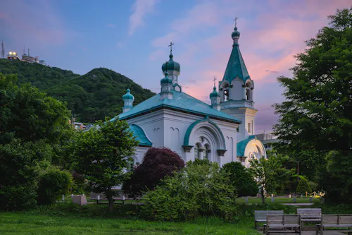 A white church with teal domes and cross-topped spires sits among lush green trees, with a backdrop of hills under a colorful pink and blue evening sky. Wooden benches are in the foreground.