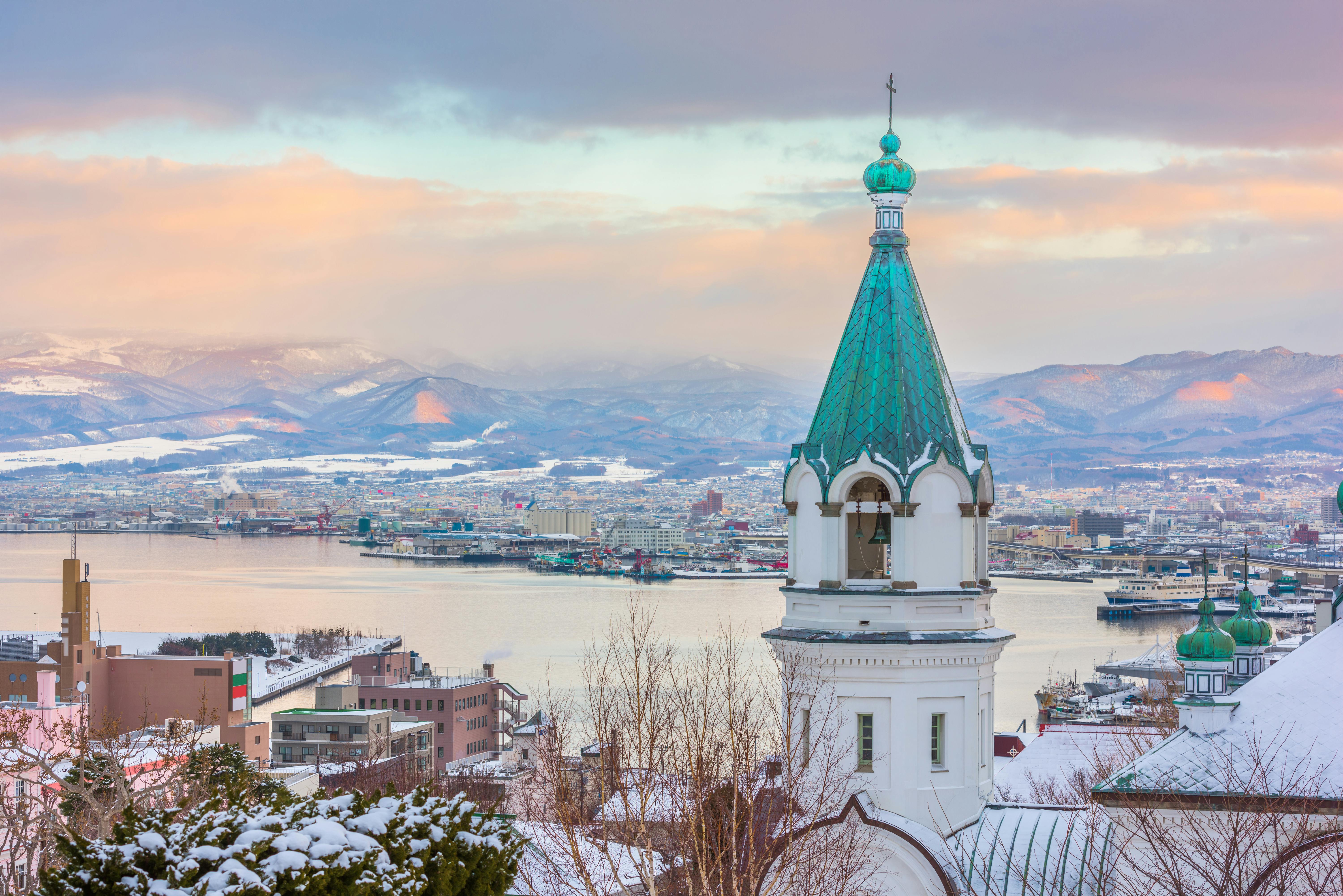 A church with a green steeple stands in the snowy city of Hakodate, Japan, overlooking the harbor and distant mountains under a pastel-colored sky at sunset.