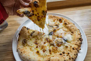 A hand lifts a slice of cheesy mushroom pizza from a white plate, with melted cheese stretching between the slice and the rest of the pizza on a wooden table.