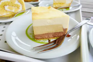 A slice of layered mousse cake with chocolate, vanilla, and yellow layers sits on a white plate with a fork beside it. The plate rests on a tray, and other dessert slices are visible in the background.