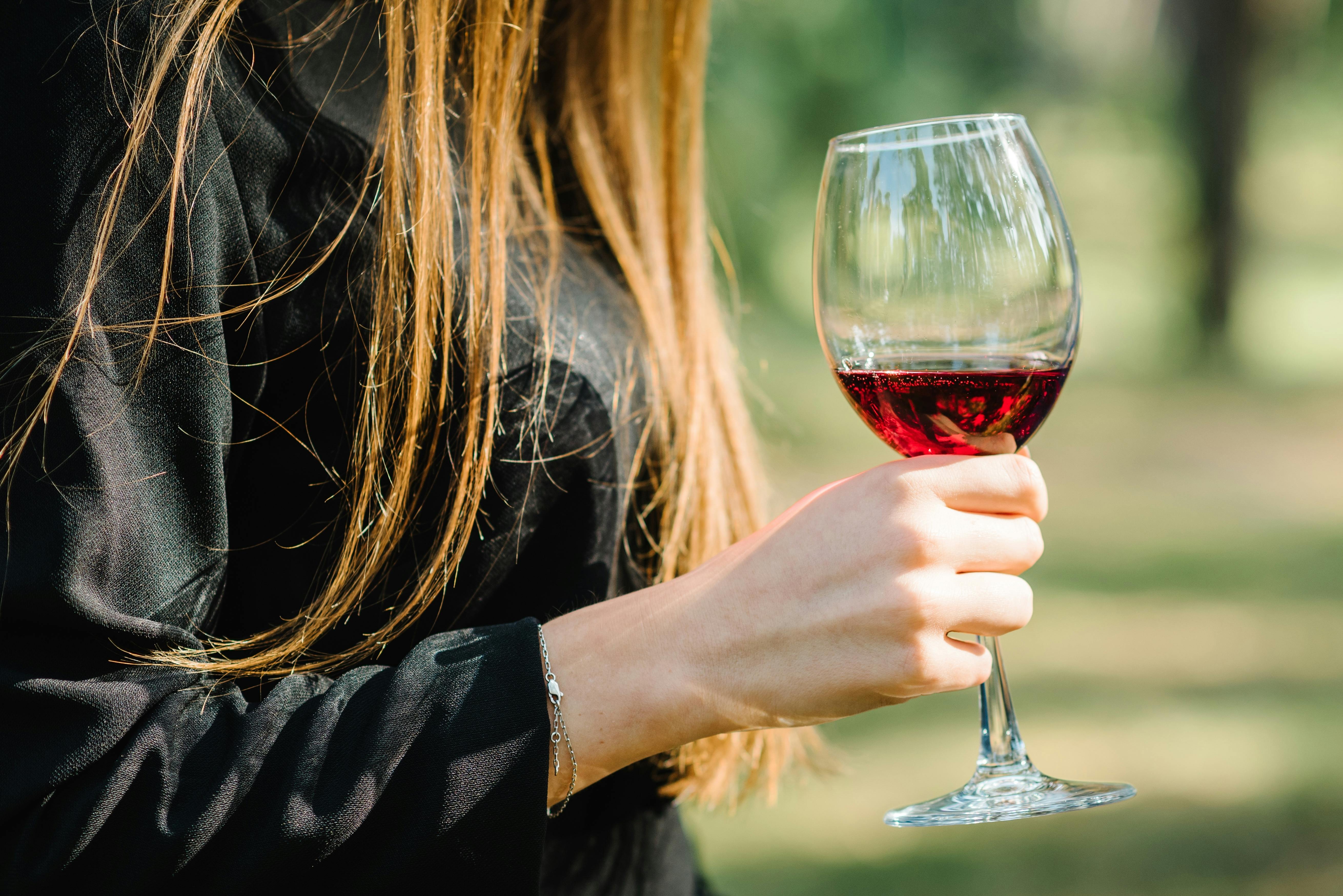 A woman with long, straight hair wearing a black top holds a glass of red wine outdoors, with sunlight illuminating her hair and a blurred green background.