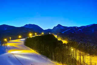 A snow-covered ski slope is brightly lit by yellow lights at dusk, surrounded by trees and mountains under a deep blue sky. The scene is tranquil, with a few distant skiers visible on the illuminated path.
