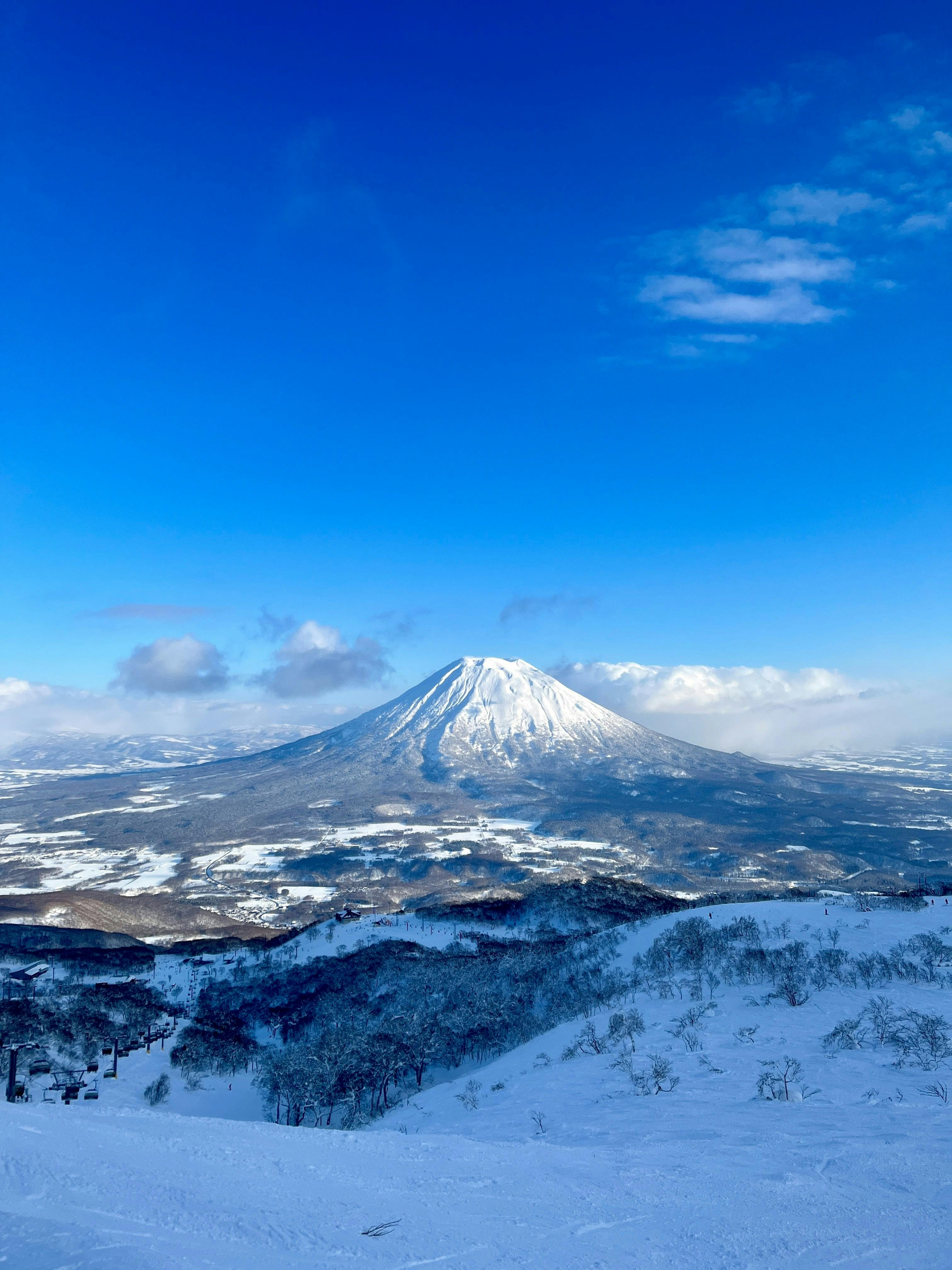 A snow-covered mountain rises under a bright blue sky with scattered clouds, surrounded by snowy fields and forests in a winter landscape.
