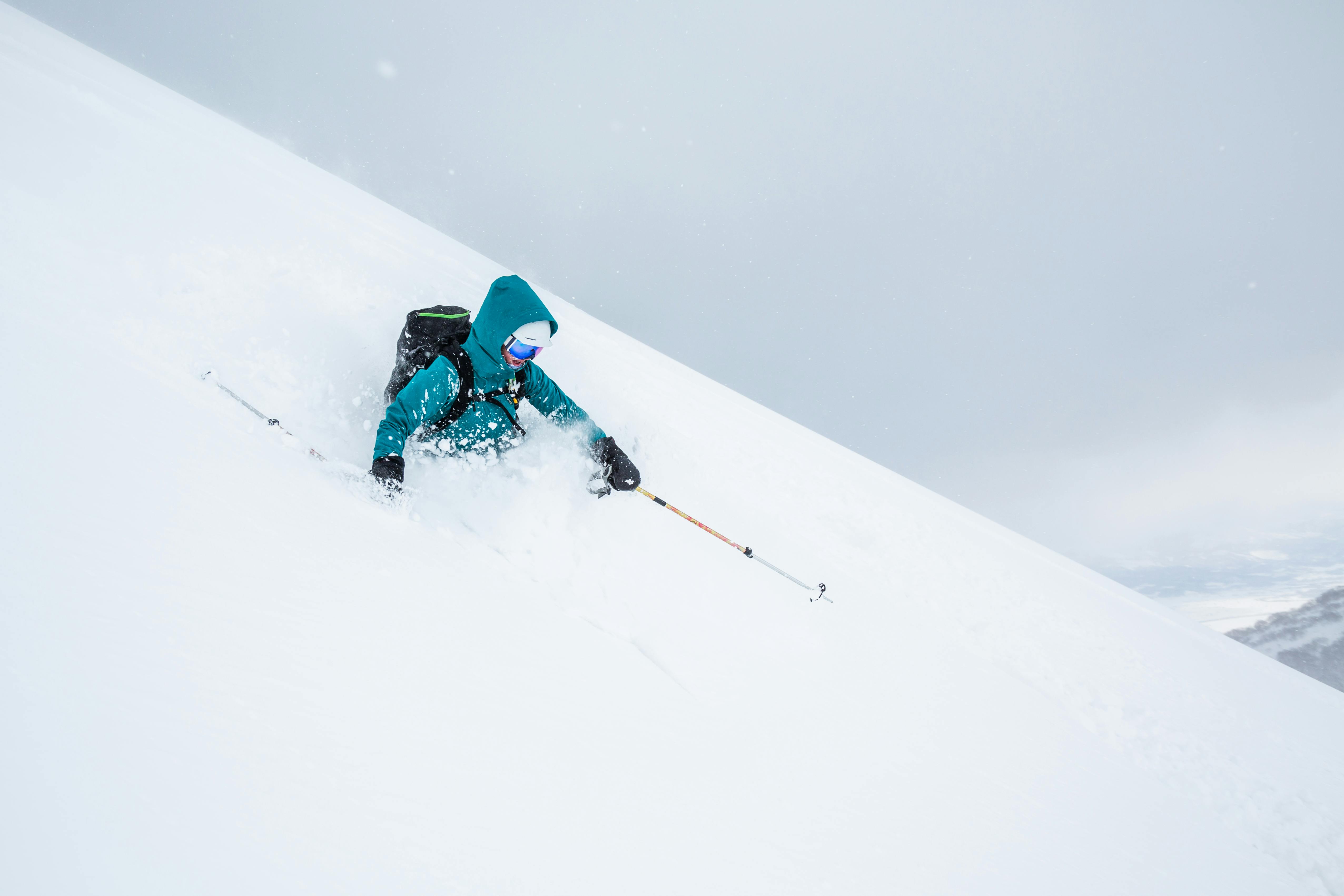 A skier wearing a teal jacket and goggles skis downhill through deep powder snow on a steep, snowy mountain slope, creating a spray of snow with ski poles extended.