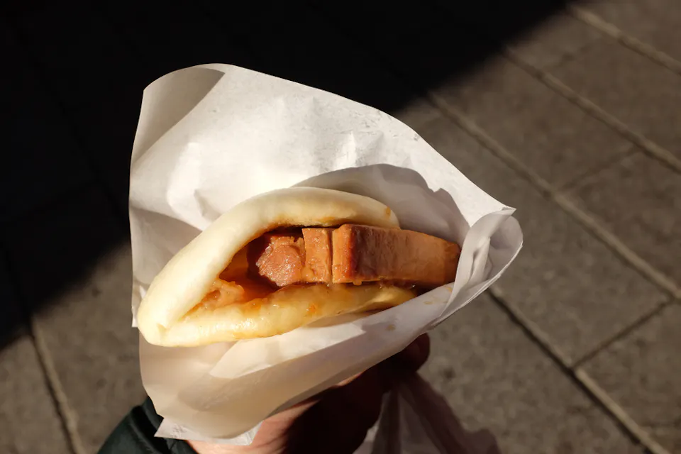 A person's hand is holding a steamed bao bun wrapped in paper. The bao is filled with a grilled slice of meat, possibly pork, along with some sauce. The background is a tiled sidewalk, slightly out of focus.