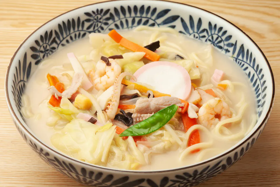 A bowl of creamy noodle soup with various toppings, including shrimp, sliced fish cakes, cabbage, carrots, green peas, corn, and other mixed vegetables. The bowl has a decorative blue floral pattern on the rim and sits on a light wooden surface.