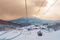 A ski resort with people skiing and a ski lift, surrounded by snowy trees and buildings, with a mountain in the background under a cloudy, golden sky.