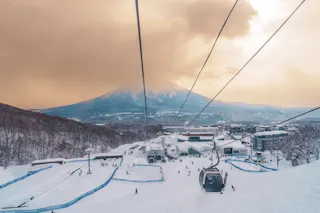 A ski resort with people skiing and a ski lift, surrounded by snowy trees and buildings, with a mountain in the background under a cloudy, golden sky.