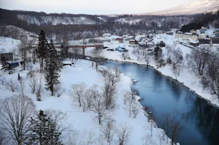 A river winds through a snowy landscape with leafless trees and a small village in the background, surrounded by snow-covered hills under a cloudy sky.