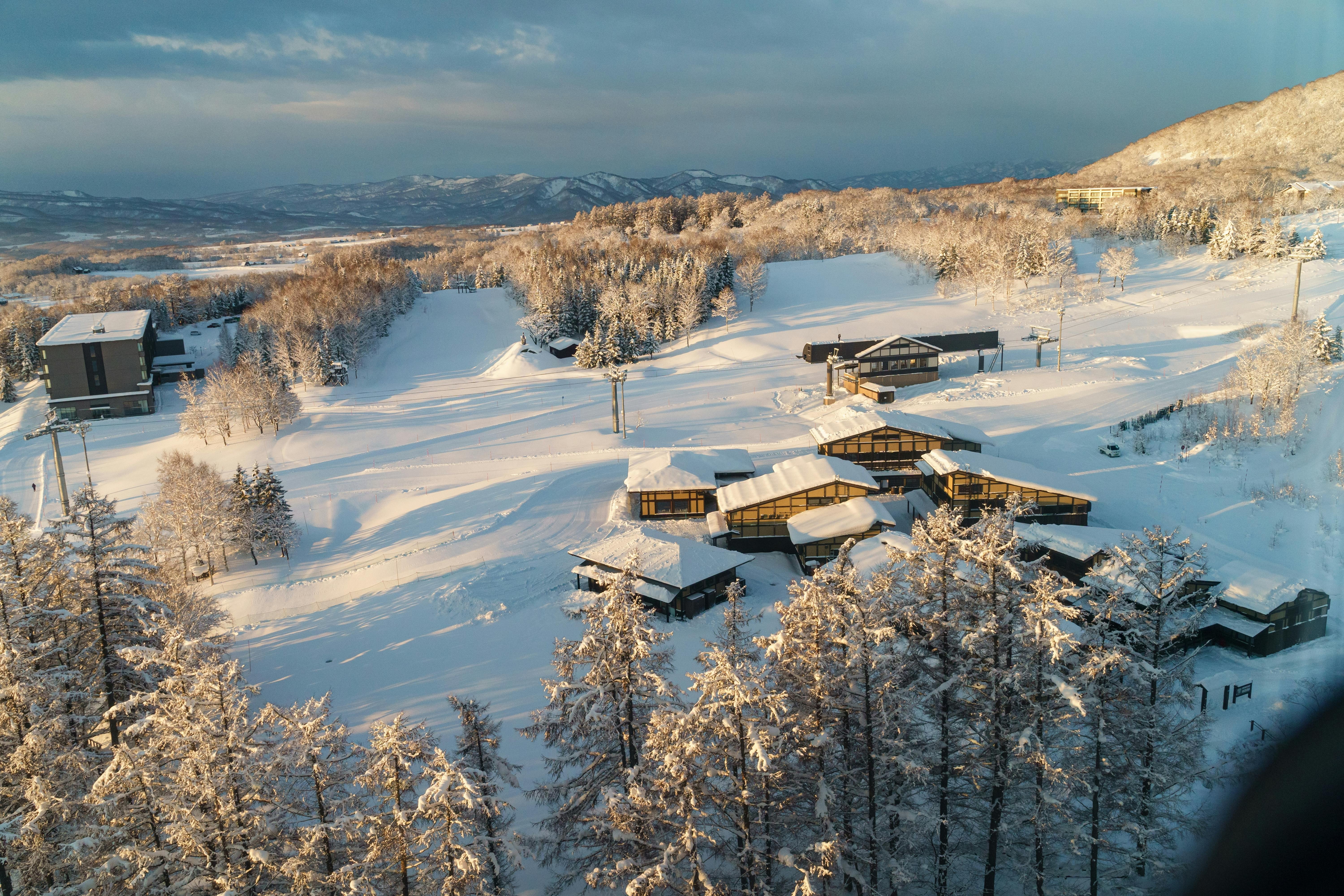 Aerial view of modern buildings surrounded by snow-covered trees and hills under a cloudy sky, capturing a serene winter landscape.
