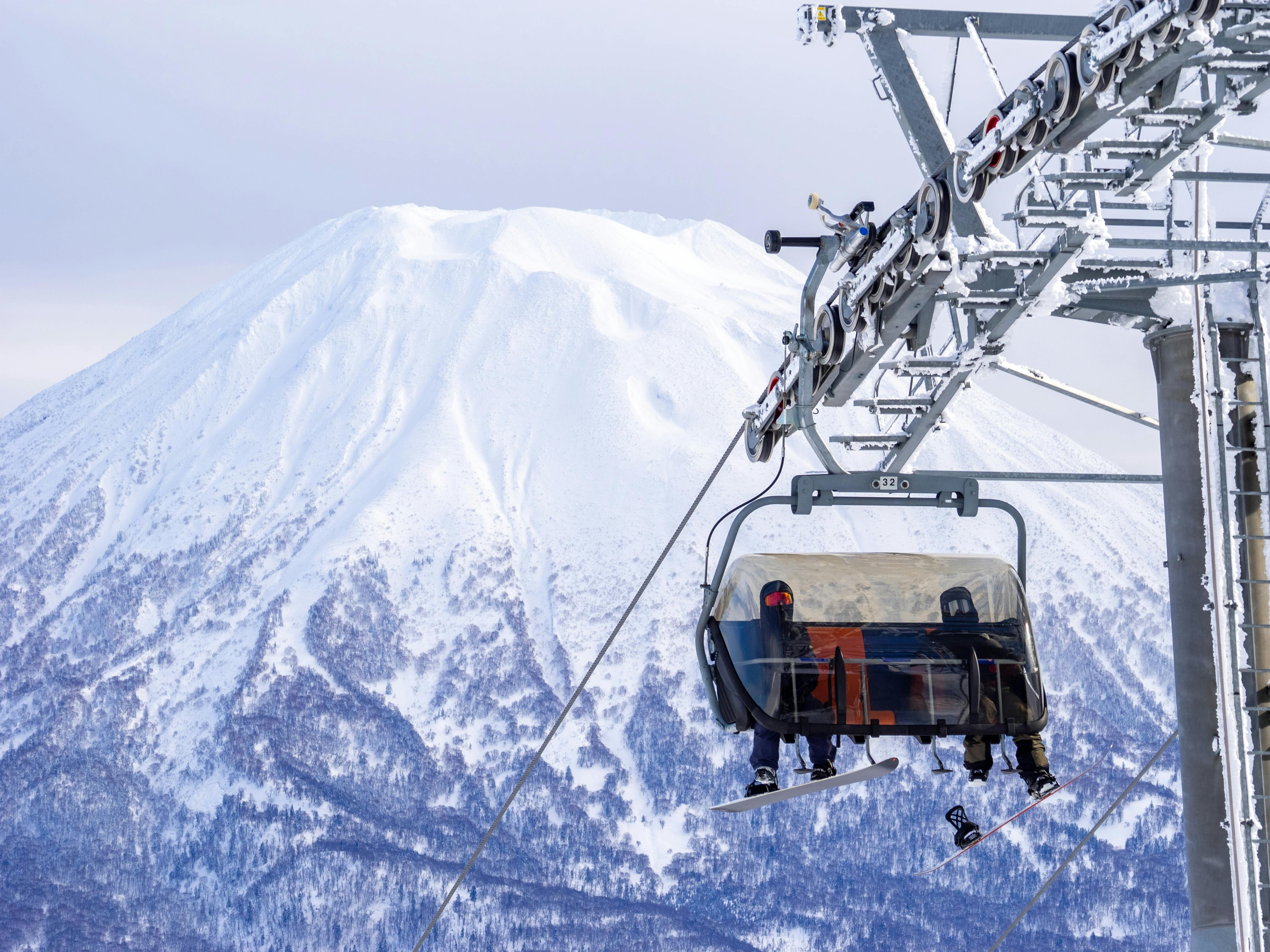 A skier rides a ski lift with a snowy mountain and trees in the background. The skier is bundled in winter clothing, and the lift is partially enclosed with a clear cover.