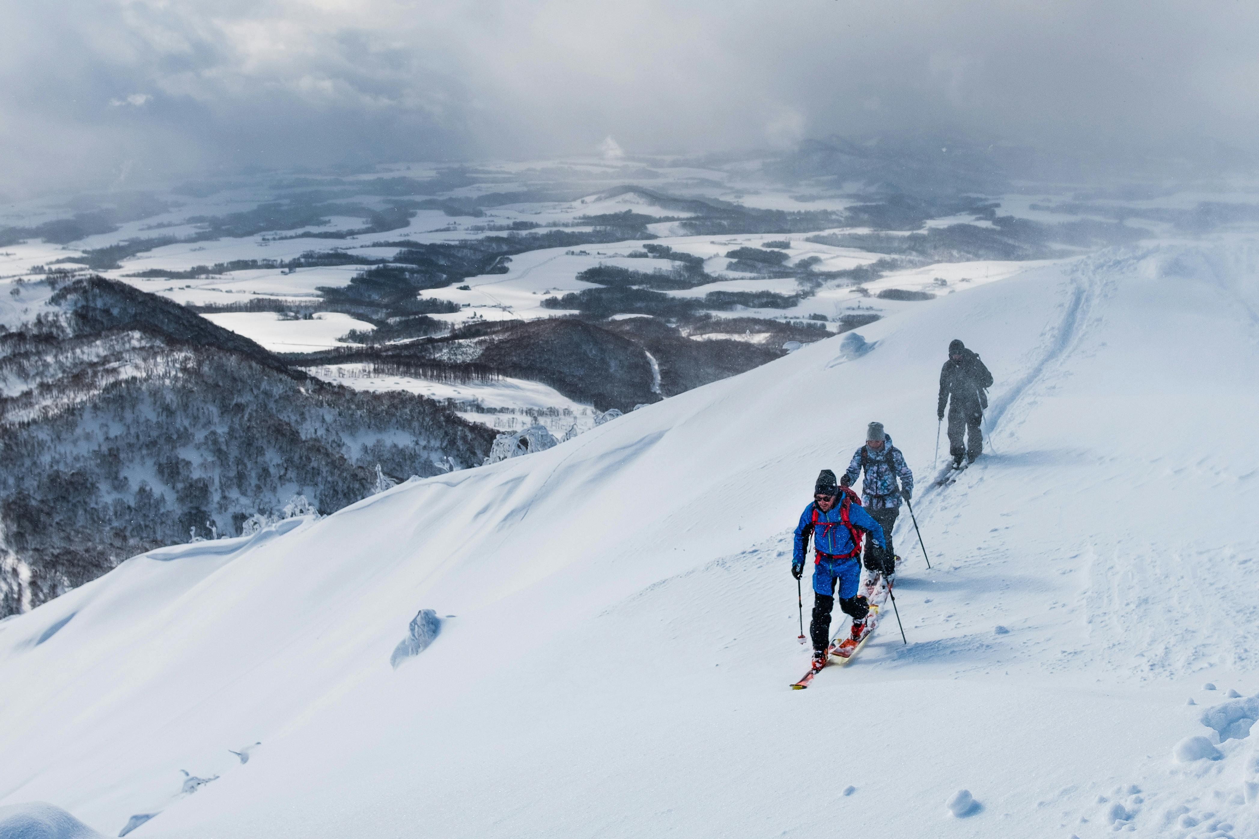 Three people in winter gear hike up a snowy mountain ridge with skis, surrounded by snow-covered hills and fields under a cloudy sky.
