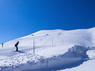 A skier is going downhill on a snowy ski slope under a clear blue sky, with ski lift poles and safety fencing visible along the slope.