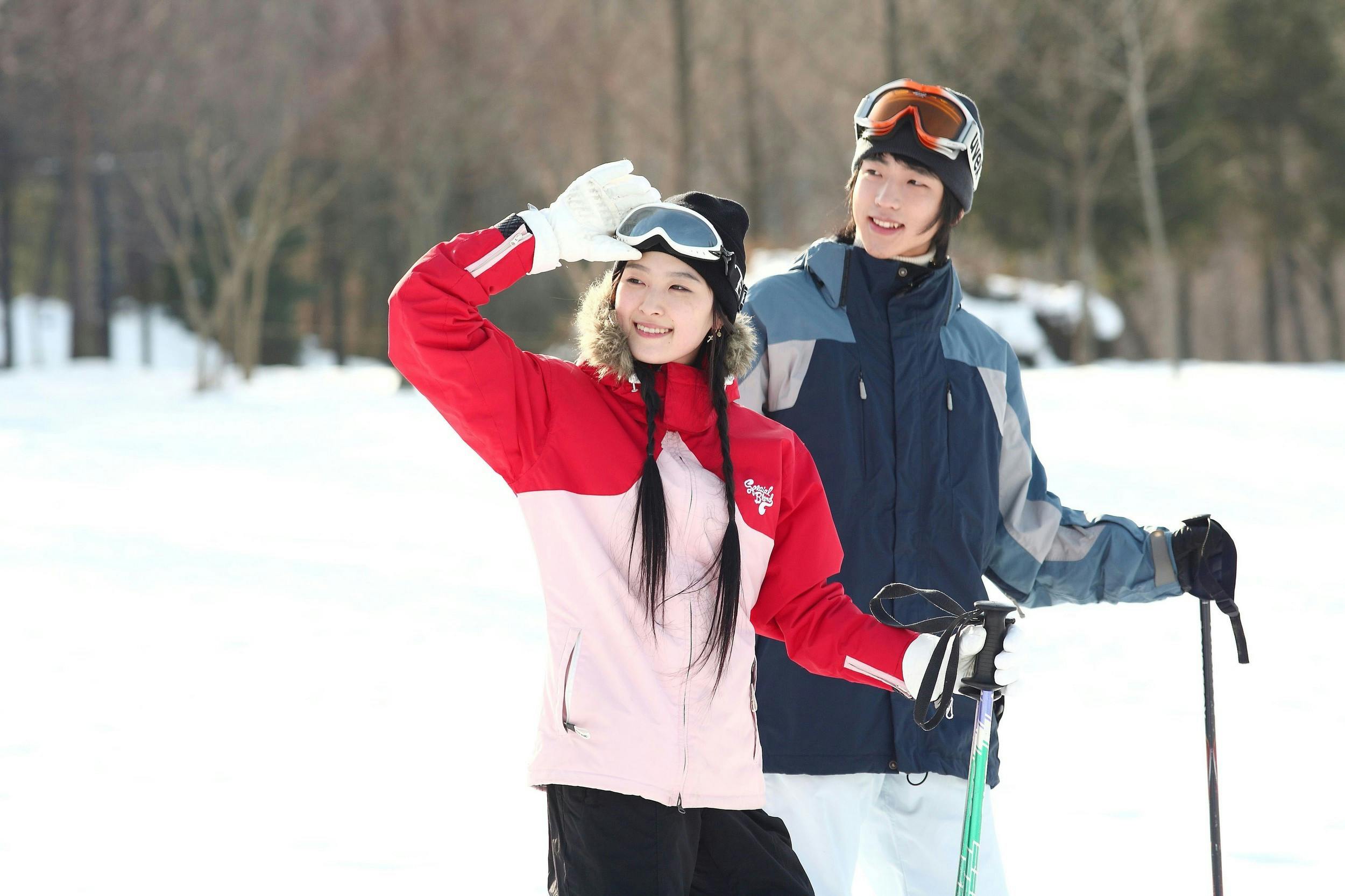 Two people dressed in winter jackets and ski gear stand on snow, smiling. One lifts a gloved hand to shield their eyes while the other holds ski poles. Trees and a snowy landscape are visible in the background.